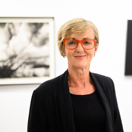 Photo of artist Jody Graham, she wears a black top and blazer and red glasses, she has short blond hair and a smile. In the background is a blurred hung black and white artwork